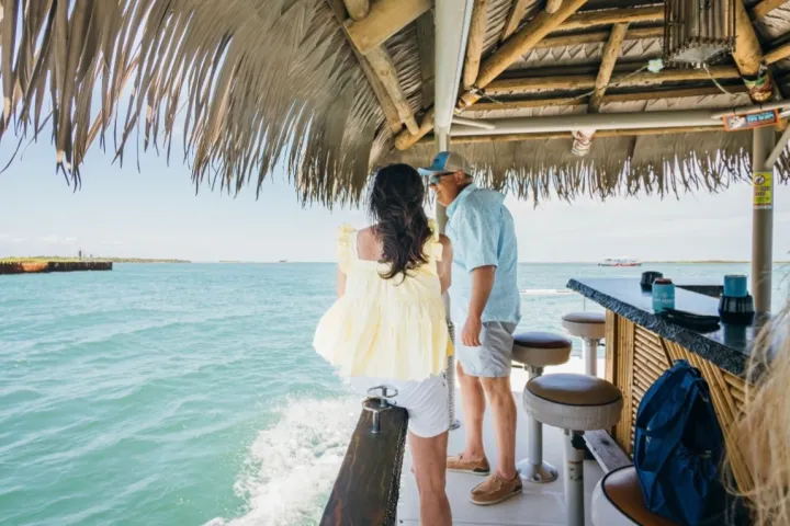 Couple on a tiki boat with thatched roof, surrounded by blue water.
