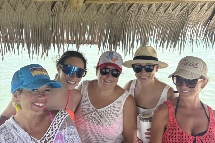 Five people smiling at a beachside tiki bar, wearing hats and sunglasses.