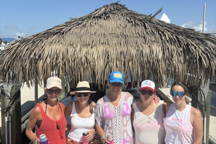Group of five people on a boat under a thatched roof, wearing casual summer attire and hats.