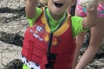Child in a red life jacket holding shells, smiling on a rocky shore.