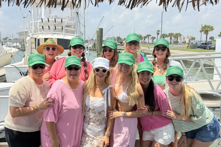 Group of people wearing green caps posing on a boat deck, with tropical hut roof and boats in the background.