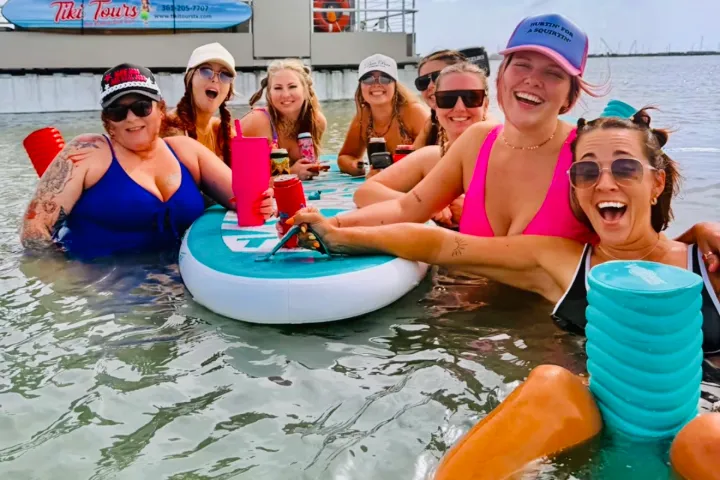 Group of women in swimwear enjoying drinks with a floating table in shallow water.