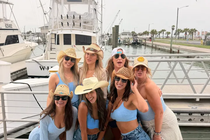 Group of seven women in summer attire and hats posing on a dock with boats in the background.