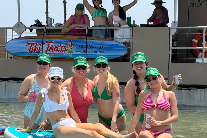 Group of women in swimsuits and hats smiling on a paddleboard near a tiki bar.