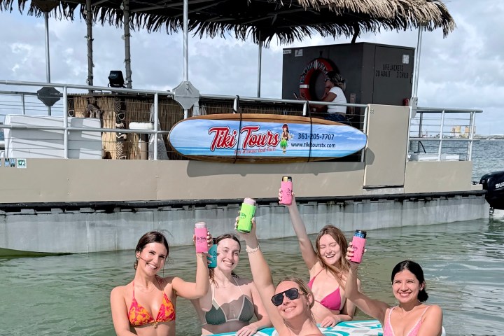 Five people in swimsuits holding drinks, standing on paddleboards near a tiki-style tour boat.