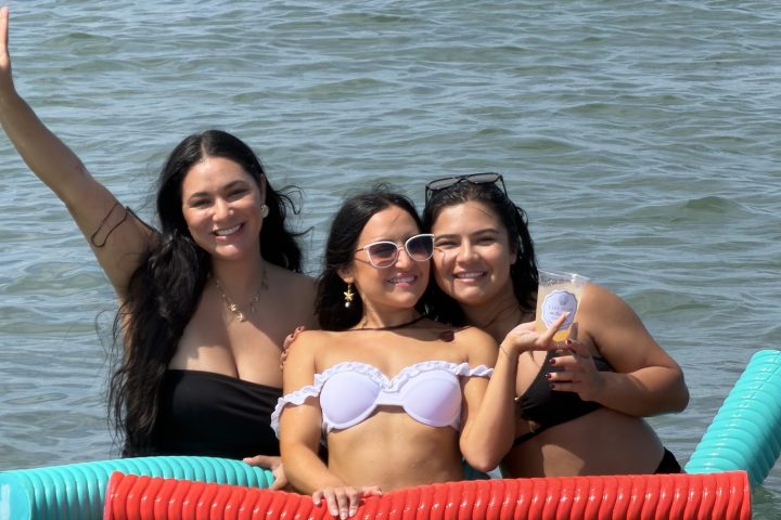 Three people on pool noodles in water, two wearing bikinis, one in black swimsuit, all smiling.