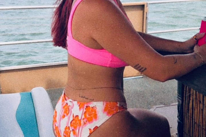 Person in pink bikini and hat sitting on boat stool by the water.
