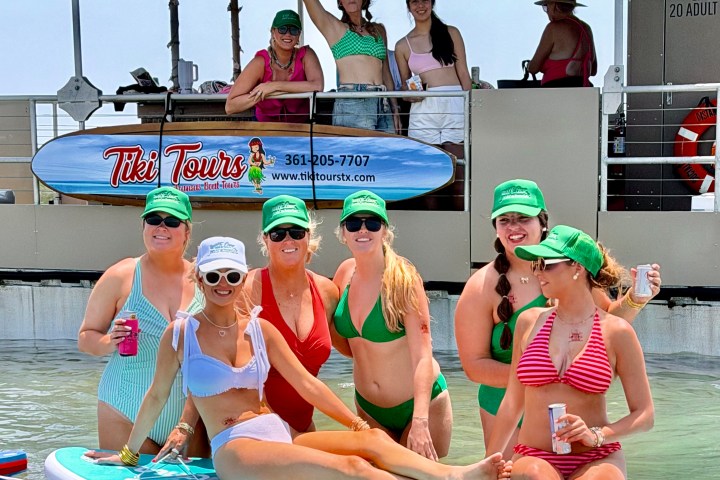 Group of women in bikinis on a paddleboard near a boat, wearing green caps and holding drinks.