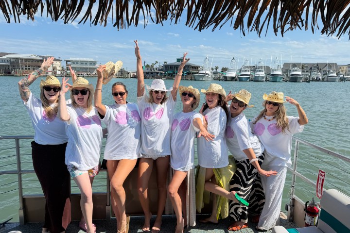 Group of women in white shirts and straw hats posing on a boat with marina background.