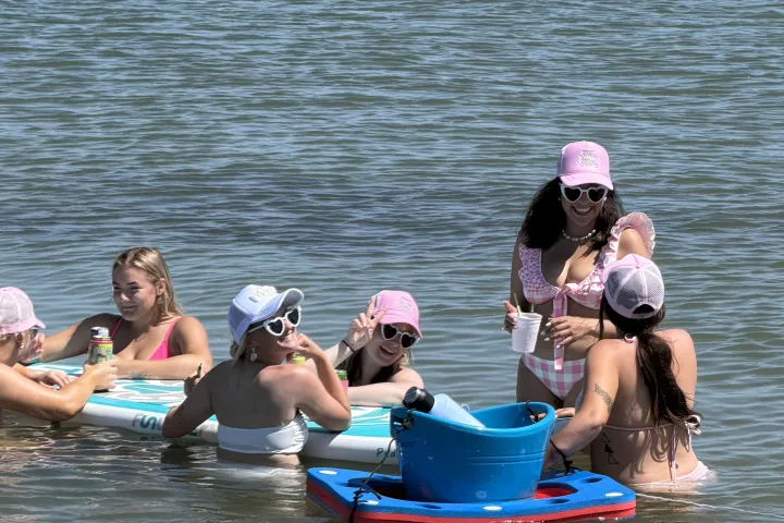 Group of people on pool floats in water, some wearing pink hats.