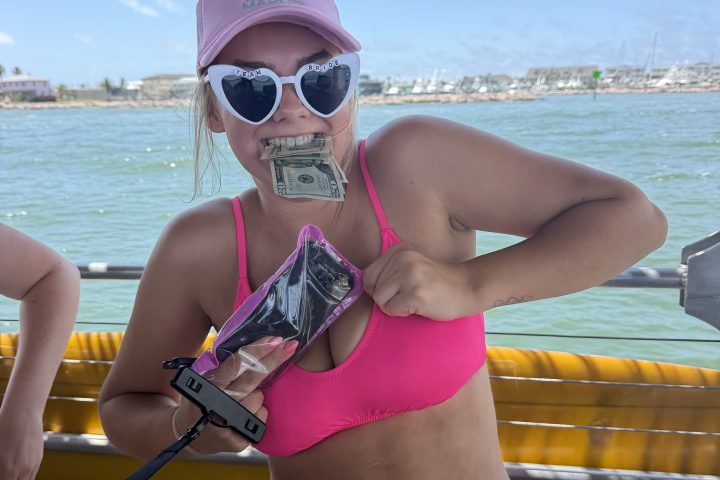 Person in pink bikini and hat holds money and phone by a waterfront.