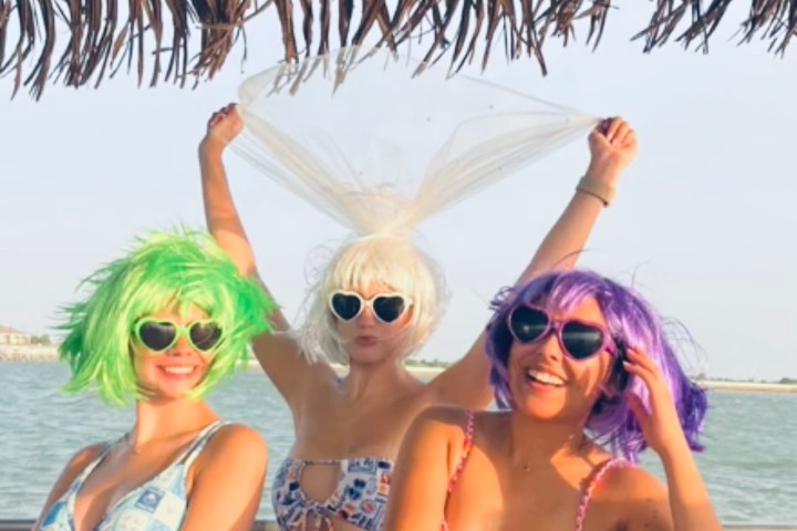 Three women in colorful wigs and bikinis pose under a thatched roof by the water.