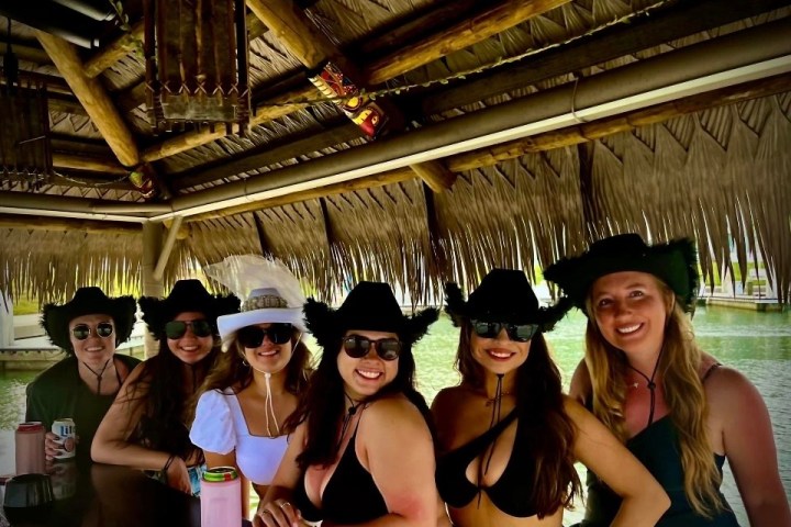 Six women in hats smiling at a tiki bar with a water backdrop.