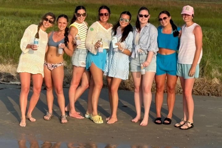 Eight women in casual beachwear standing on sand, holding drinks, with greenery in the background.