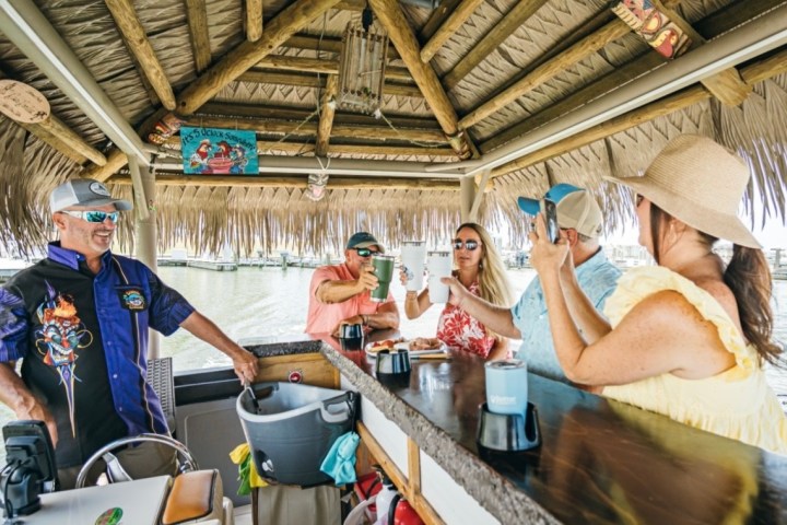 Group of people enjoying drinks at a tiki bar on a boat with a thatched roof.