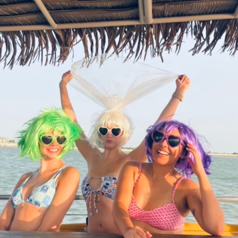 Three people in colorful wigs and bikinis pose playfully under a thatched roof with water in the background.