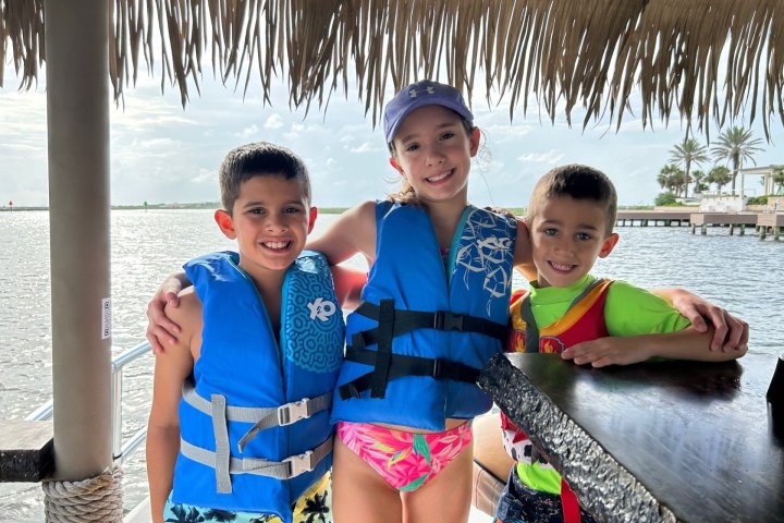 Three children in life jackets smiling under a thatched roof by the water.