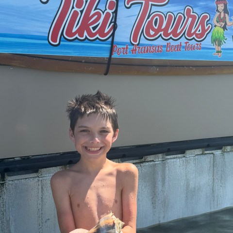 Smiling boy holds large seashell, standing near Tiki Tours sign by the water.