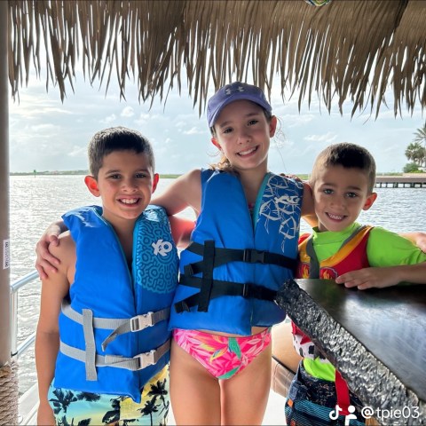 Three children in life jackets on a boat under a thatched roof.
