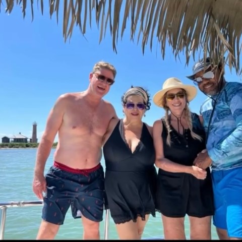 Four people in swimwear posing on a boat under a thatched roof, with water in the background.