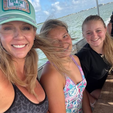 Three people with a beach background, two smiling and one with hair wind-swept, under a straw canopy.
